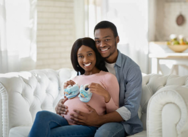 Pregnant woman and her husband sitting together in their living room, smiling and holding tiny baby shoes, symbolizing excitement and anticipation while choosing a delivery method.