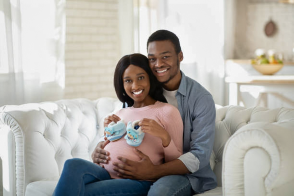 Pregnant woman and her husband sitting together in their living room, smiling and holding tiny baby shoes, symbolizing excitement and anticipation while choosing a delivery method.
